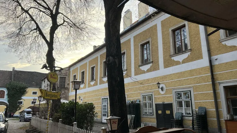 Historisches Gebäude mit gelber Fassade, alten Bäumen und einem Außenbereich, der als Biergarten dient.
