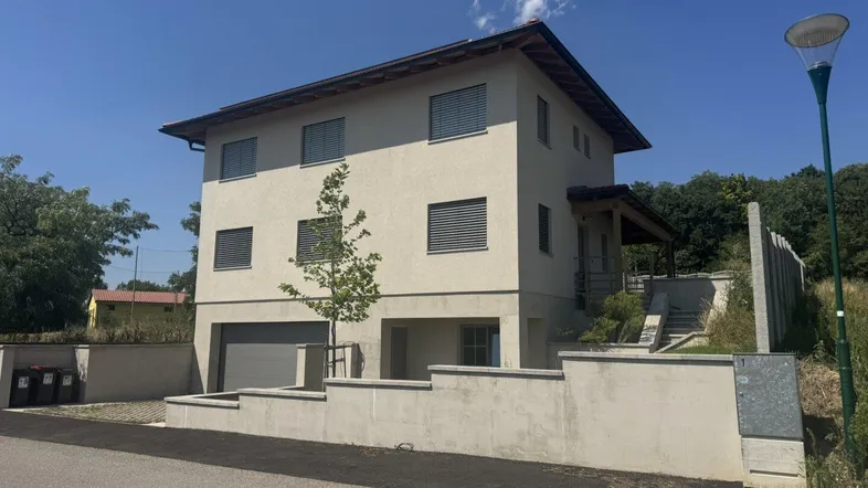 Modernes Einfamilienhaus mit heller Fassade, Rollläden und integrierter Garage unter blauem Himmel.