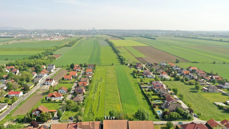 Luftaufnahme einer ländlichen Wohnsiedlung umgeben von weiten grünen Feldern mit Fernblick auf die Stadt.