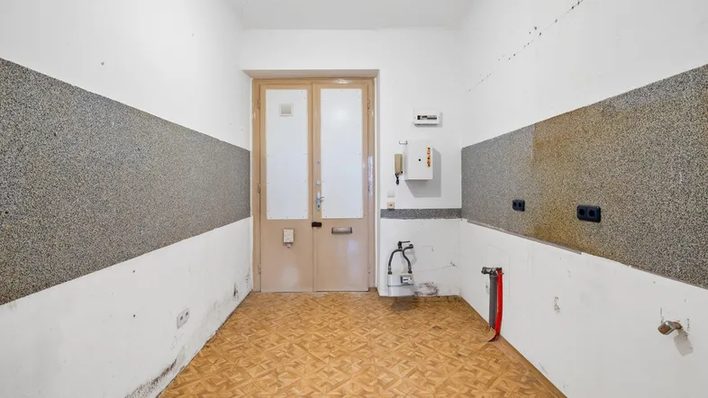 Leere Küche mit alten Vinylfliesen und sichtbaren Anschlüssen für Gas und Wasser.