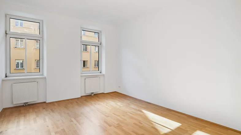 Helles Zimmer mit Holzboden und zwei Fenstern, die viel Tageslicht hereinlassen.