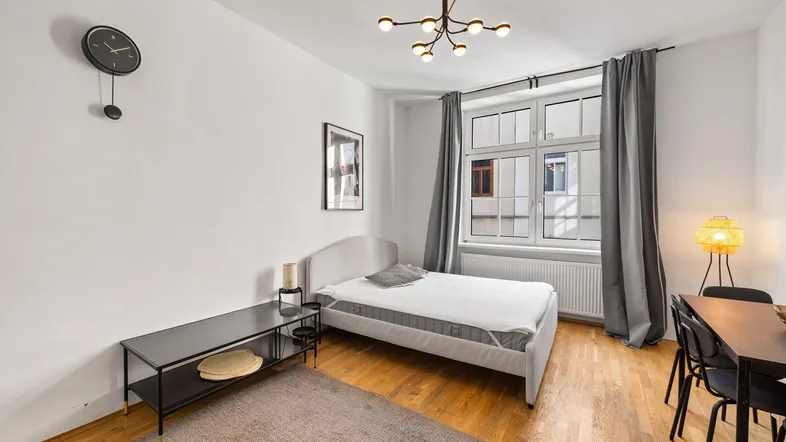 Helles Schlafzimmer mit Holzboden, großem Fenster und modernem Kronleuchter, ideal für Einzelpersonen.