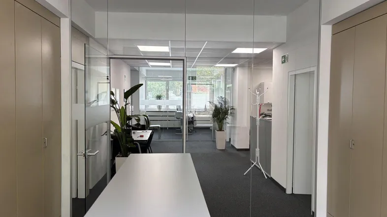 Moderner Büroraum mit Glaswänden, Teppichboden und integrierten Schränken, bietet viel natürliches Licht.