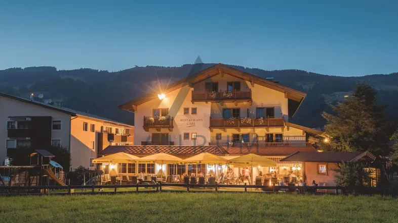 Abendaufnahme eines traditionellen Hotels mit beleuchtetem Restaurantbereich und Bergblick bei Dämmerung.