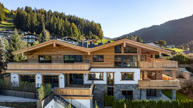 Moderne Chalet-Architektur mit Holzfassade, Steinakzenten und mehreren Balkonen vor bewaldeter Bergkulisse.