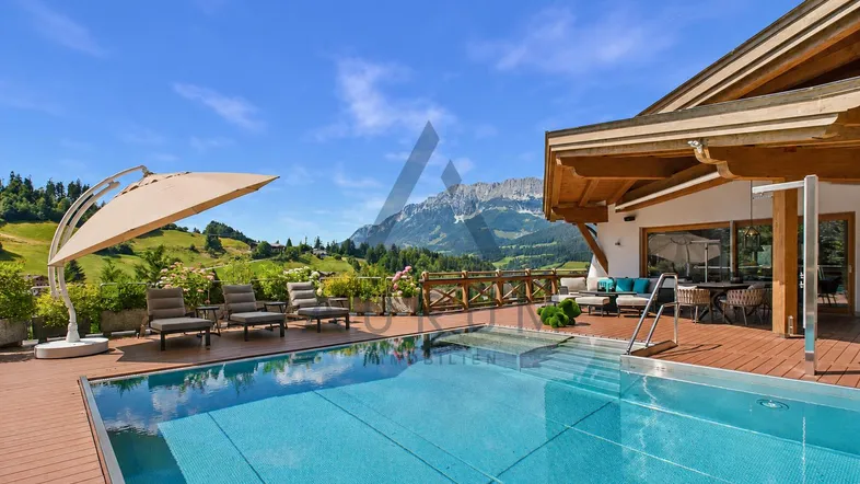 Große Sonnenterrasse mit Infinity-Pool, Liegestühlen und atemberaubendem Bergblick unter blauem Himmel.