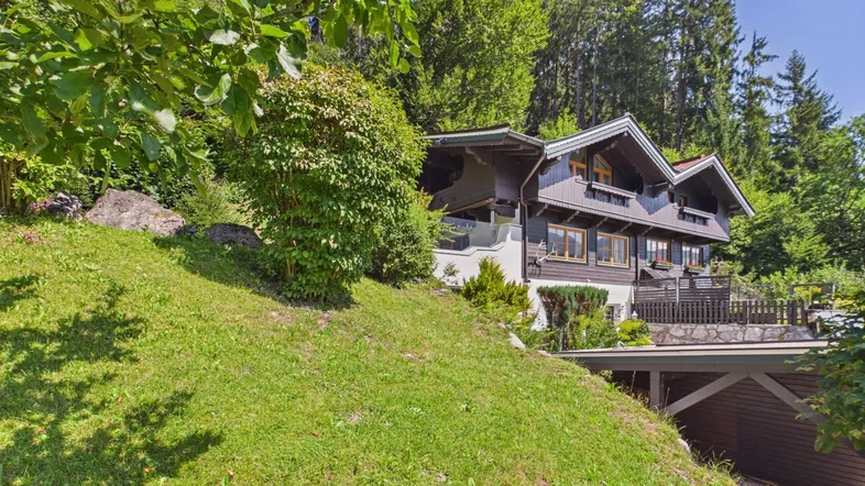 Traditionelles Haus am Hang mit viel Grün und einem Carport im Vordergrund.