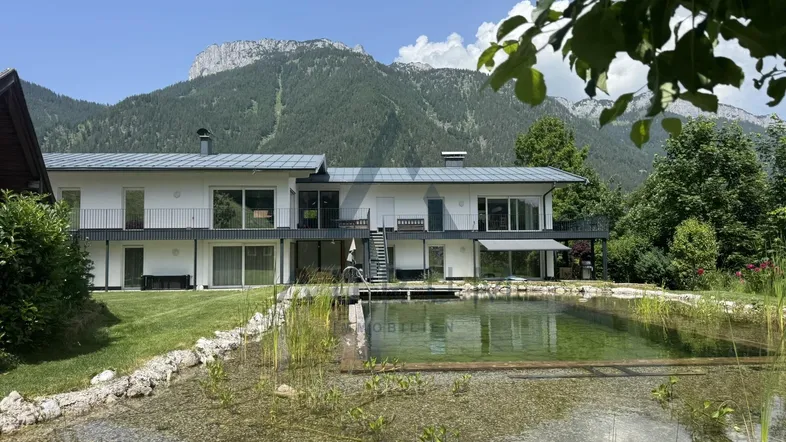 Weitläufiger Garten mit Naturteich und moderner Architektur im Hintergrund, umgeben von Bergen.