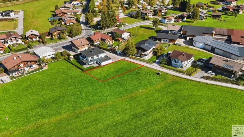 Luftaufnahme eines grünen Baugrundstücks mit roter Markierung, umgeben von Wohnhäusern und Natur.