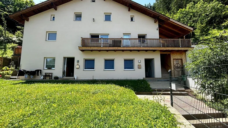Traditionelles Wohnhaus mit Holzbalustrade und gepflegtem Vorgarten in sonniger Hanglage mit Bergblick.