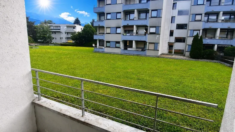 Blick vom Balkon auf eine grüne Rasenfläche und ein Mehrfamilienhaus mit Bergblick.