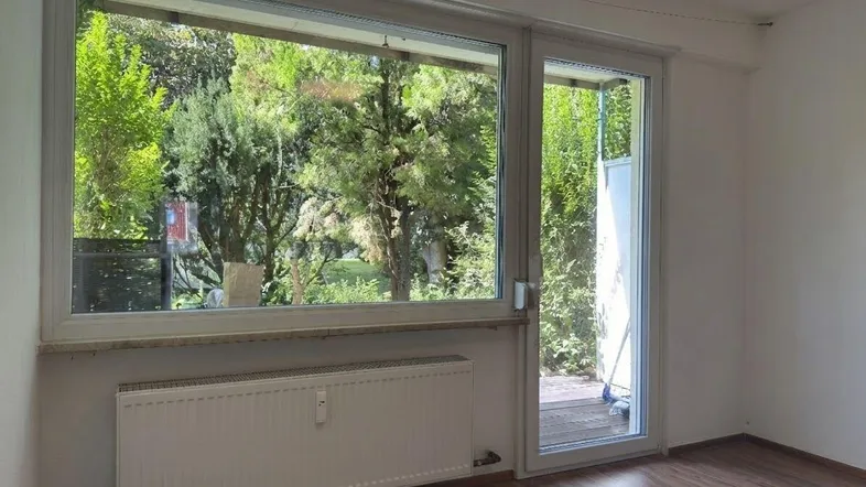 Helles Zimmer mit Holzboden und großem Fenster, das Zugang zu einem Außenbereich bietet.
