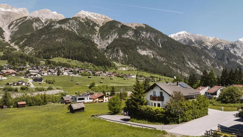 Luftaufnahme eines Hauses in einer malerischen Berglandschaft mit grünen Wiesen und schneebedeckten Gipfeln.