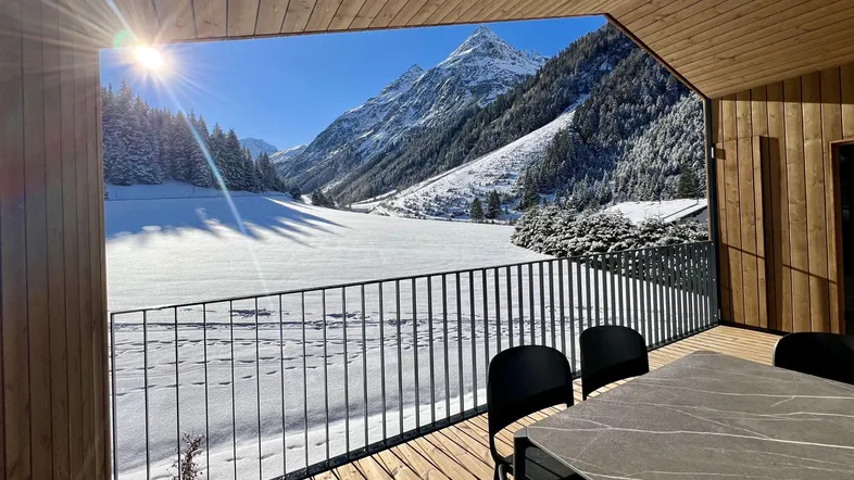 Sonniger Balkon mit Holzboden, Esstisch und atemberaubendem Bergblick auf die verschneite Landschaft.