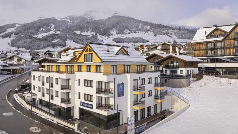 Modernes Apartmentgebäude in den verschneiten Bergen mit traditionellen Holzelementen und Balkonen.
