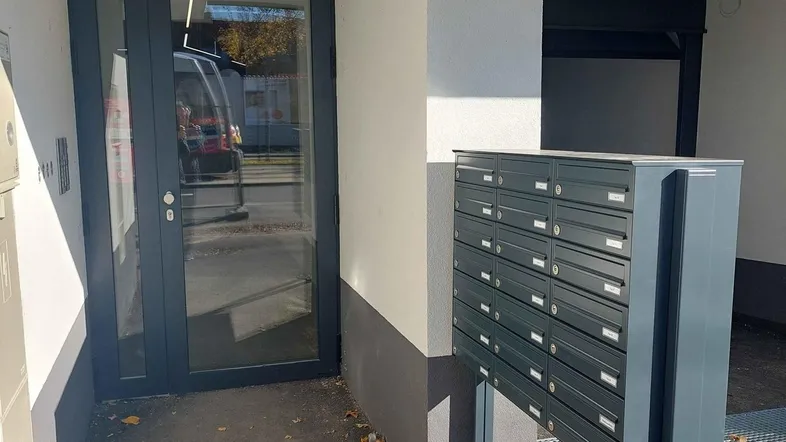 Eingangsbereich eines modernen Gebäudes mit Briefkästen und Glastür an einem sonnigen Tag.