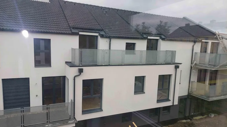 Neubau-Fassade mit modernen Balkonen und dunklem Ziegeldach, aufgenommen von einem Fenster aus.