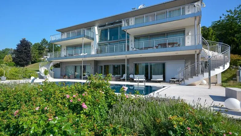 Moderne Villa mit großzügigem Poolbereich, mehreren Balkonen und üppiger Gartenanlage unter blauem Himmel.