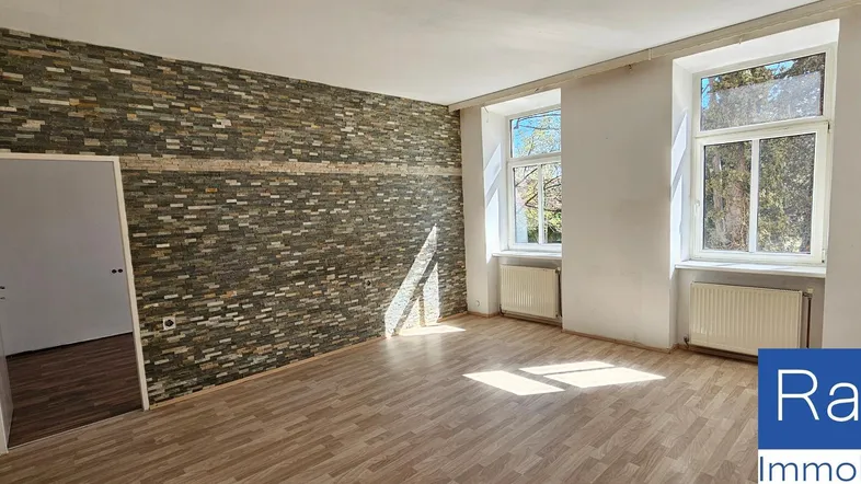 Heller Wohnraum mit markanter Steinwand, Holzboden und großen Fenstern, die viel Tageslicht hereinlassen.