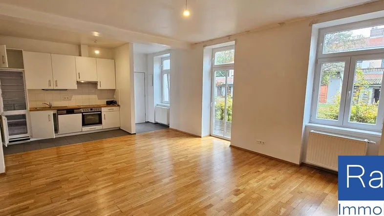 Helles Wohn- und Esszimmer mit integrierter Küche, Parkettboden und großen Fenstern.