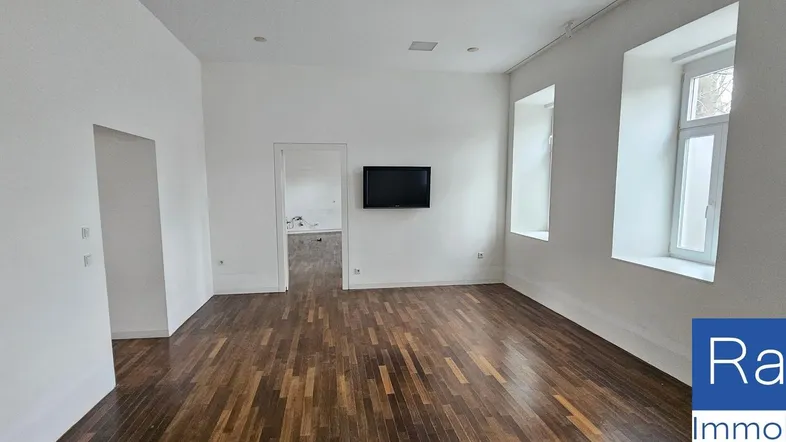 Helles, leeres Zimmer mit dunklem Parkettboden und einem an der Wand montierten Fernseher.