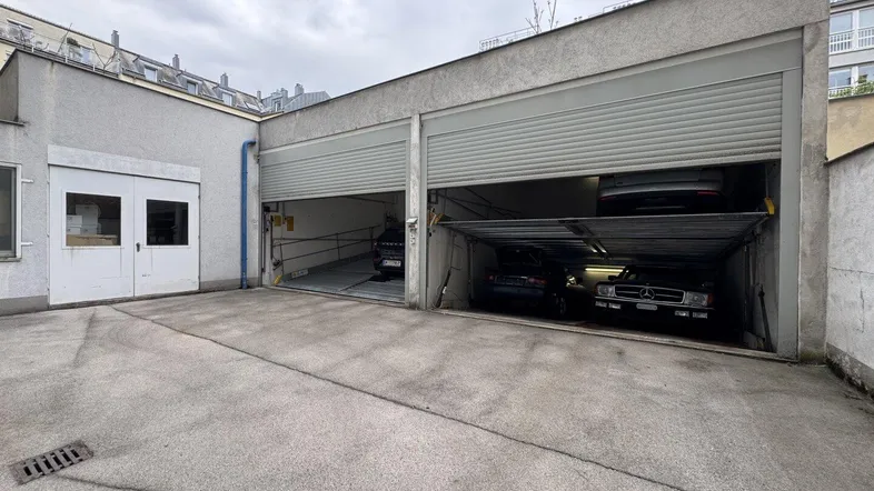 Außenbereich der Garage mit zwei Stellplätzen und angrenzendem Nebengebäude auf einem betonierten Vorplatz.