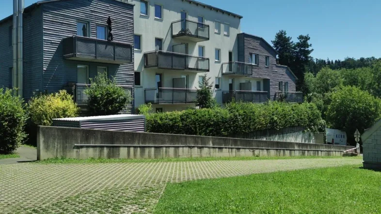 Mehrfamilienhaus mit mehreren Balkonen und grüner Umgebung unter blauem Himmel.