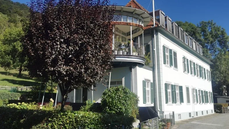 Weißes Gebäude mit markantem Rundbalkon und grünen Fensterläden in hügeliger Landschaft.