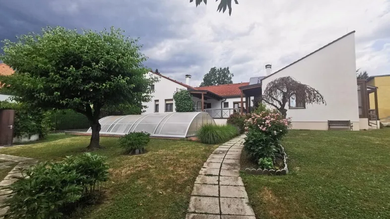 Großer Garten mit gepflegtem Rasen, Bäumen und einem überdachten Poolbereich unter bewölktem Himmel.