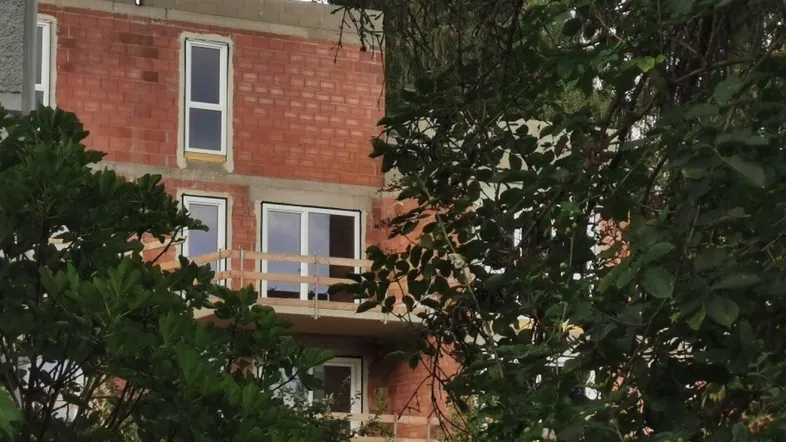 Neubau in Rohbauweise mit sichtbaren Ziegelwänden und provisorischen Balkongeländern, umgeben von Bäumen.