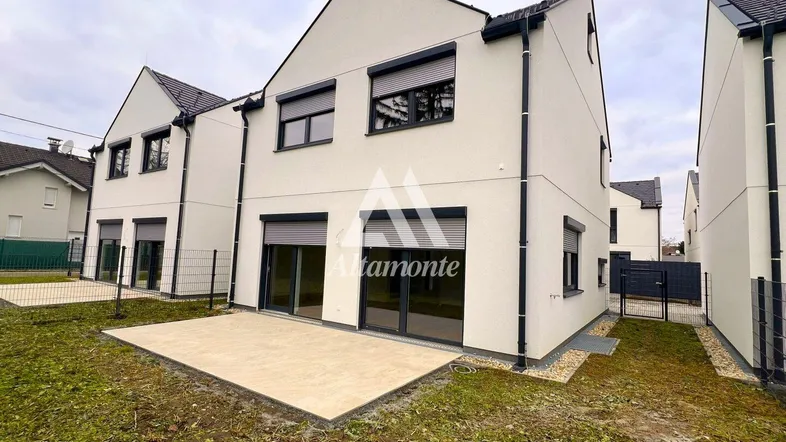 Neubau-Reihenhaus mit moderner Fassade, Rollläden und einer gepflasterten Terrasse im Gartenbereich.