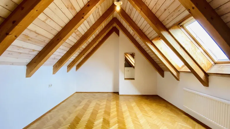 Helles Dachzimmer mit sichtbaren Holzbalken, Holzdecke und Fischgrätparkettboden.