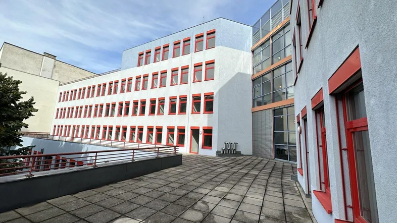 Große gepflasterte Terrasse vor einem modernen Bürogebäude mit roten Fensterrahmen unter blauem Himmel.