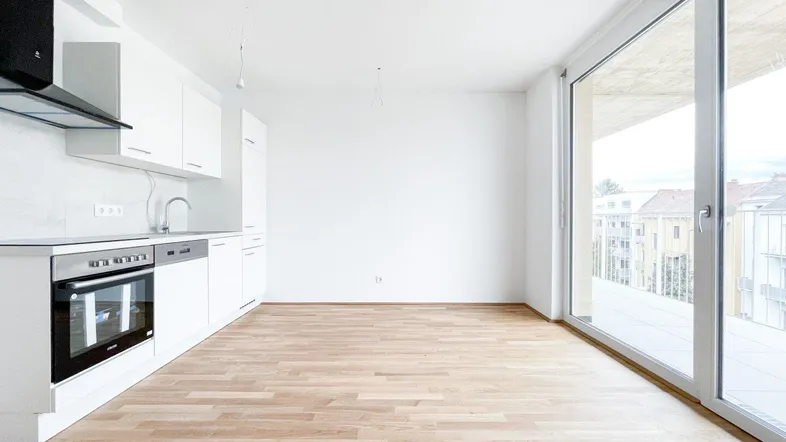 Heller offener Wohnbereich mit integrierter Küche, Holzboden und Zugang zu einem großen Balkon.