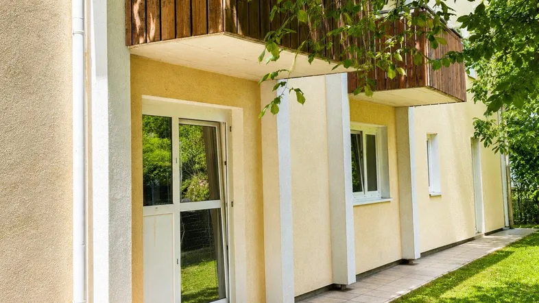 Fassade des Gebäudes mit gelbem Putz, Holzbalkon und Zugang zum Gartenbereich.