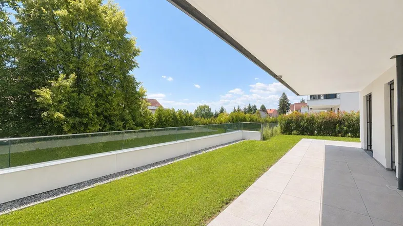 Moderner Gartenbereich mit gepflegtem Rasen, heller Terrasse und Glasgeländer unter blauem Himmel.
