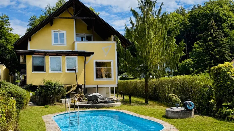 Gelbes Haus mit Satteldach und einem gepflegten Garten, der einen Swimmingpool und alte Bäume umfasst, umgeben von grüner Natur.