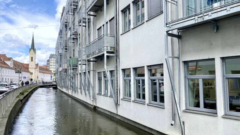 Gebäude mit Balkonen entlang eines Kanals, mit Kirchturm im Hintergrund unter bewölktem Himmel.