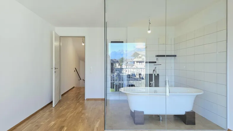 Modernes Badezimmer mit freistehender Badewanne in Glasabtrennung und Holzboden, angrenzend an einen Wohnbereich.