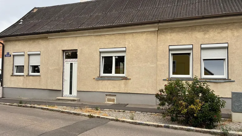 Einfaches Einfamilienhaus mit beiger Fassade, Rollläden und einem kleinen Busch vor dem Eingang.
