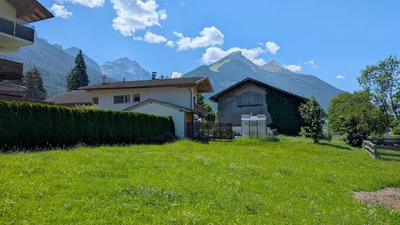 Blick auf ein Haus und eine Scheune mit gepflegtem Rasen und majestätischen Bergen im Hintergrund unter blauem Himmel.