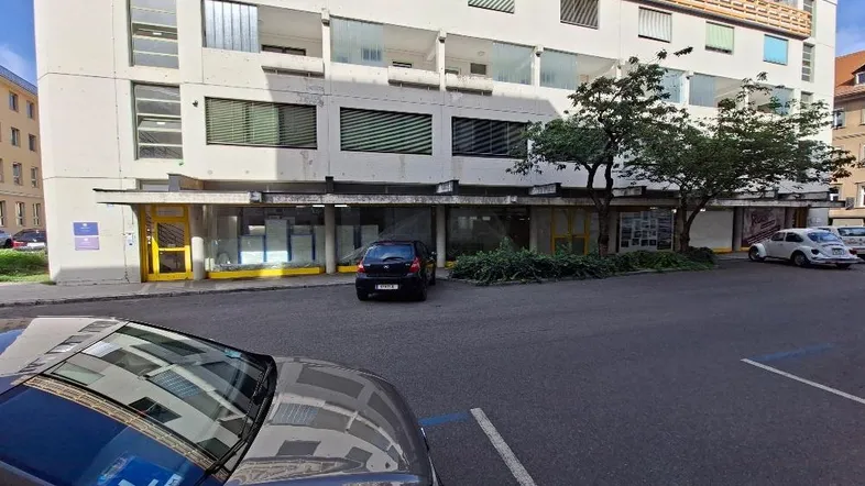 Gebäudeansicht mit mehreren Etagen, Geschäftsräumen im Erdgeschoss und Parkplätzen davor.