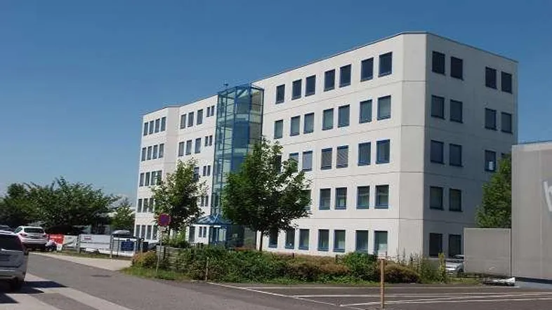 Modernes Bürogebäude mit heller Fassade, vielen Fenstern und einem gläsernen Aufzugsschacht.