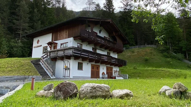 Traditionelles Alpenhaus mit Holzbalkonen und grüner Umgebung, eingebettet in eine bewaldete Berglandschaft.