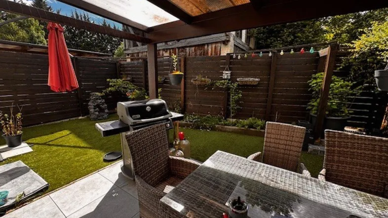 Überdachte Terrasse mit Essbereich, Grill und Kunstrasenfläche, umgeben von einem Holzzaun.