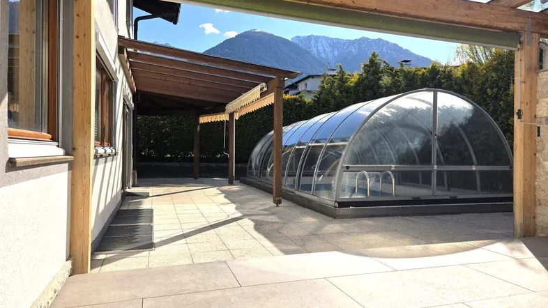 Große geflieste Terrasse mit überdachtem Pool, Holzpergola und beeindruckendem Bergblick.