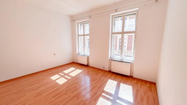 Helles, leeres Zimmer mit Laminatboden und zwei großen Fenstern, die viel Tageslicht hereinlassen.