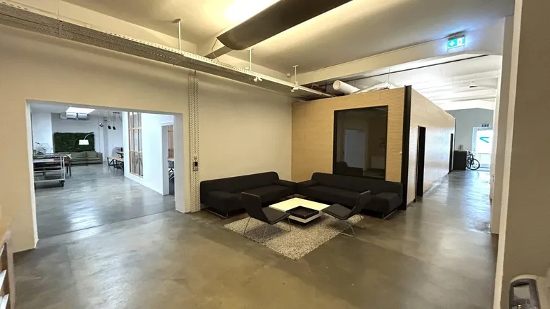 Großzügiger offener Bürobereich mit Lounge-Ecke, Betonboden und sichtbaren Deckeninstallationen.