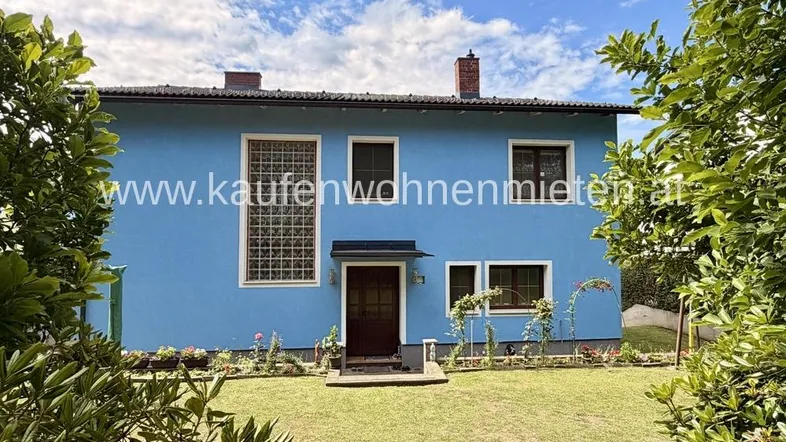Blaues Einfamilienhaus mit gepflegtem Vorgarten und grüner Umgebung unter blauem Himmel.