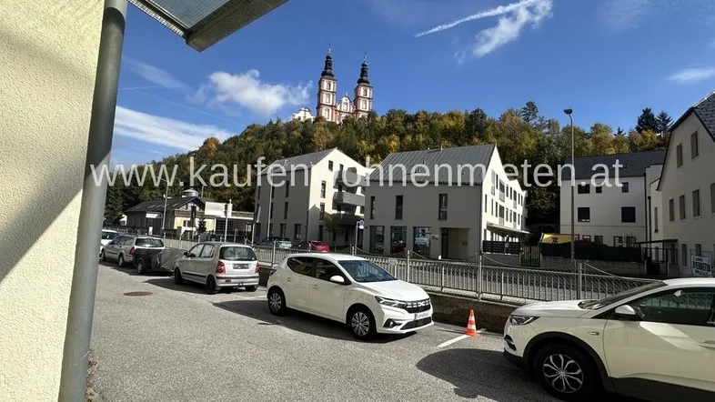 Blick auf eine Straße mit geparkten Autos, modernen Gebäuden und einer Kirche auf einem bewaldeten Hügel im Hintergrund.